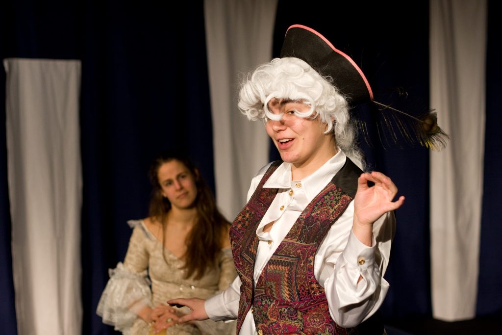 Im Vordergrund des Bildes ist Carolin Falke als Leutnant Riccaud zu sehen, im Hintergrund steht Lilith Rüschenpöhler als Minna von Barnhelm und lauscht dem Monolog Leutnant Riccaud. Sie stehen auf der Bühne vor einem blau-weißem Vorhang.