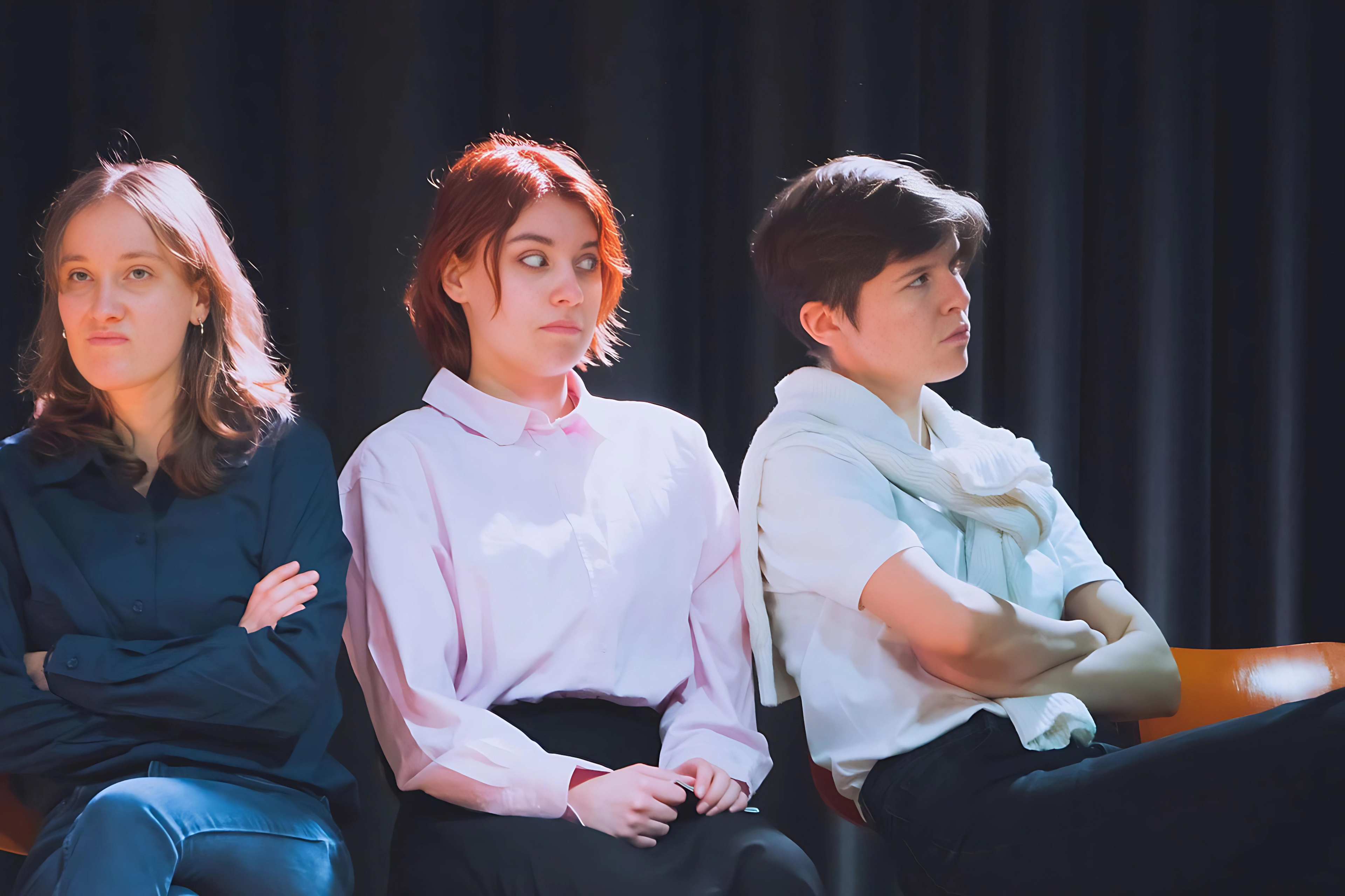 Die drei Hauptdarstellerinnen des Theaterstückes "Kunst" während der Probe. Sie sitzen gemeinsam auf der Bühne vor einem schwarzen Vorhang.