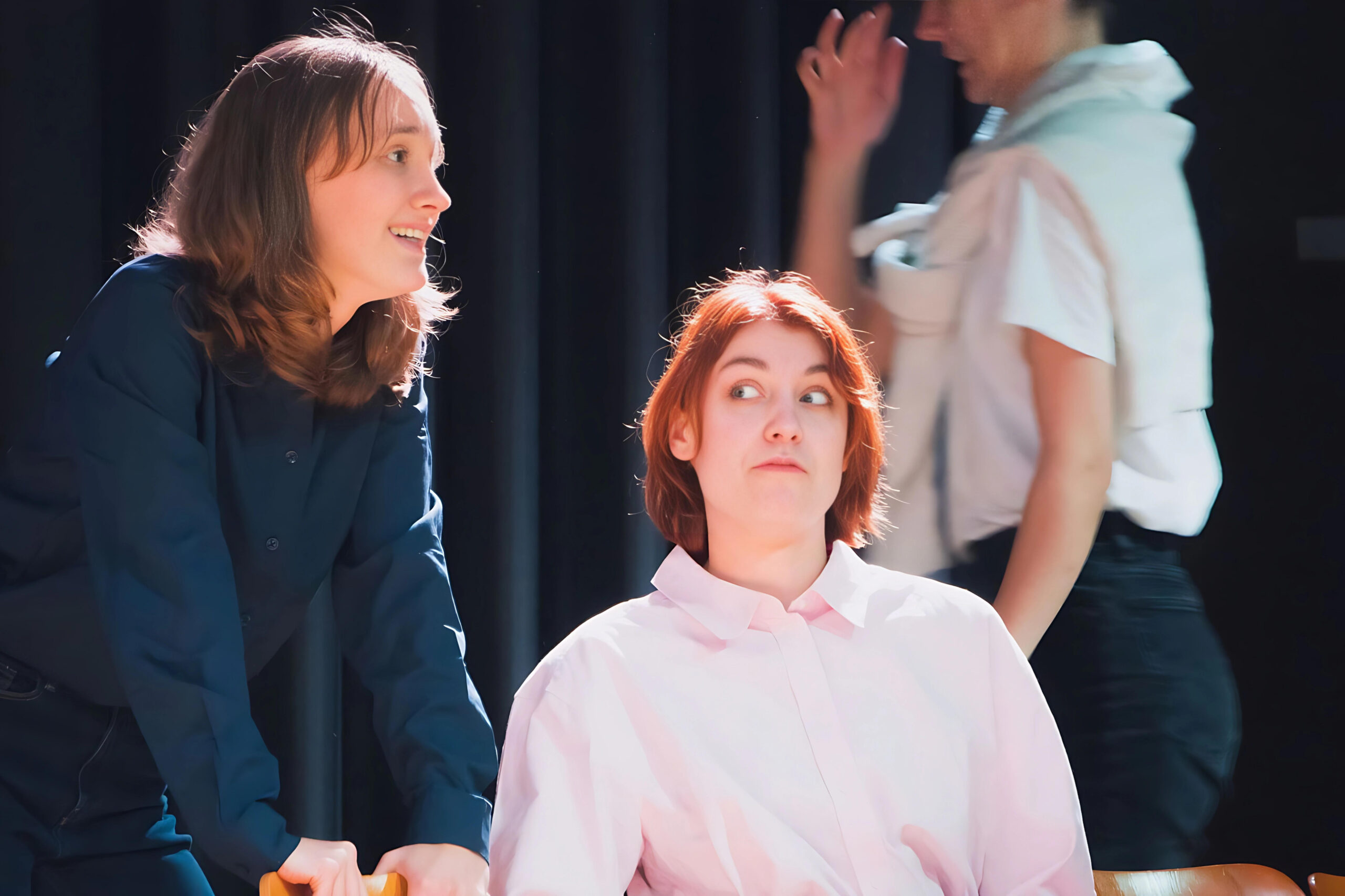 Die drei Hauptdarstellerinnen des Theaterstückes "Kunst" während der Probe. Die Schauspielerin links im Bild steht, die Schauspielerin in der Bildmitte sitzt und die Schauspielerin rechts im Bild geht im Hintergrund an ihnen vorbei.