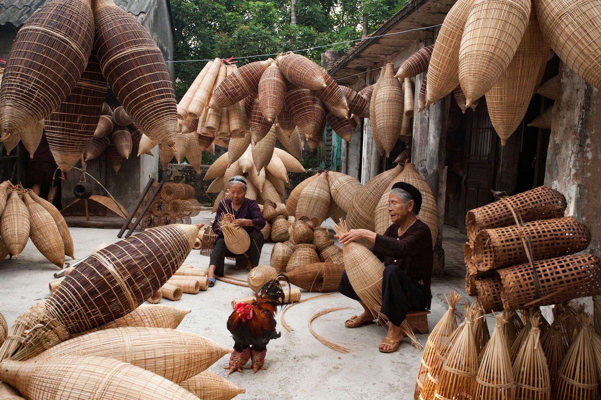 Szene aus Vietnam. Zwei Frauen flechten auf dem Boden sitzend Körbe aus Holz; um sie herum hängen viele Körbe.